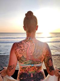 Rear view of shirtless tattooed woman at beach against sky during sunset