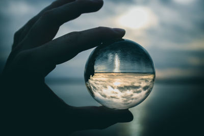 Close-up of hand holding crystal ball against sky during sunset
