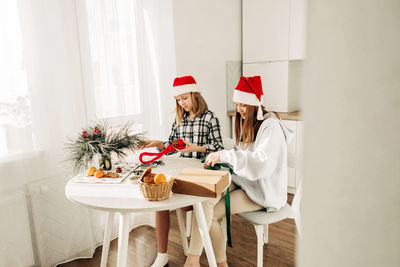Two cute girls in santa claus hats are sitting at the kitchen table and packing sweet gifts