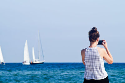 Rear view of woman photographing with mobile phone while standing by sea against sky