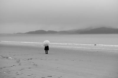 Rear view of person with umbrella standing at beach against cloudy sky