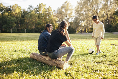 Sisters sitting on log while boy playing with ball at park during sunny day