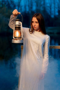 Young woman with illuminated oil lamp standing in forest at dusk