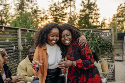 Cheerful woman with arm around female friend holding drink glass at dinner party