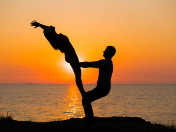 Silhouette couple exercising at beach against sky during sunset