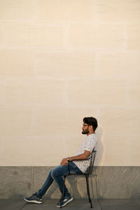 Side view of man sitting on chair against wall