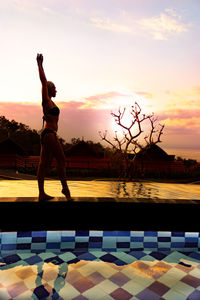 Woman standing by swimming pool against sky during sunset