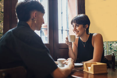 Happy young woman holding coffee cup talking with boyfriend in cafe