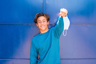 Portrait of smiling teenage boy holding mask while standing against wall