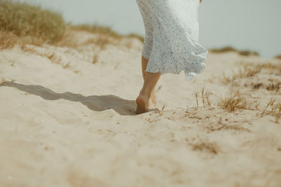 Back view of crop unrecognizable barefoot female in white summer dress walking alone on sandy beach in sunny summer day in spain