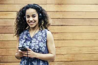 Portrait of smiling young woman listening to music from mobile phone while standing against wooden wall