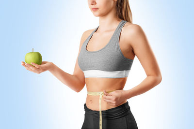 Midsection of young woman holding apple while measuring waist against white background