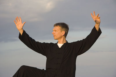 Mid adult man with arms raised looking away while standing against cloudy sky