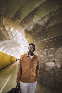 Smiling young man carrying bag while standing with hand in pocket at subway tunnel