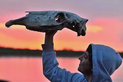 Close-up of man holding animal skull against lake during sunset