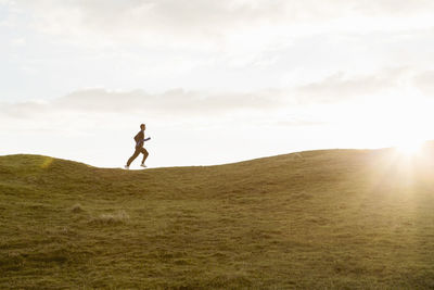 Distant view of man jogging on grassy hill against sky during summer