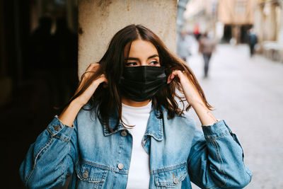 Confident young female in trendy denim clothes and black protective mask for coronavirus prevention looking away while standing near stone column on city street