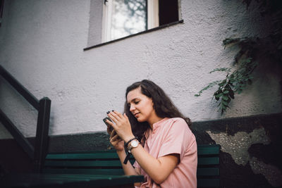 Young woman using mobile phone while sitting on bench outside house