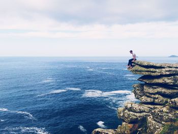 Man sitting at the edge of cliff over sea against sky