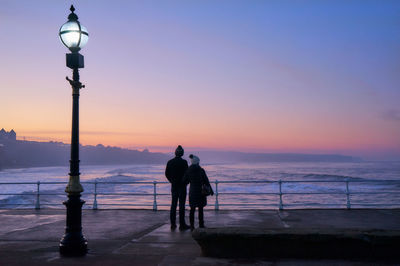 Silhouette men on street by sea against sky at sunset