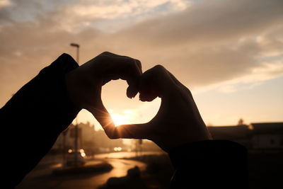 Hand holding heart shape against sky during sunset