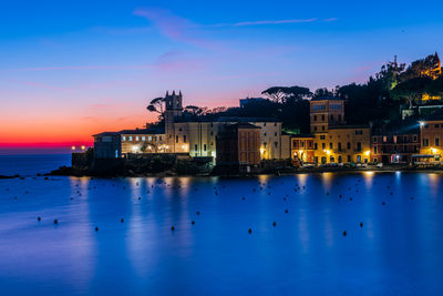 Illuminated buildings by sea against sky at sunset