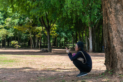 Side view of woman crouching by tree trunk and using smart phone at park