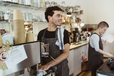 Smiling owner wearing apron looking away while standing near coffee maker at cafe