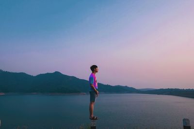 Full length side view of man standing on wooden post in lake against sky during sunset