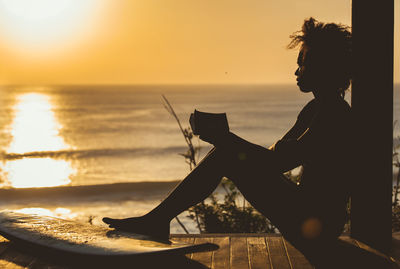 Side view of silhouette woman holding book against sea sky during sunset