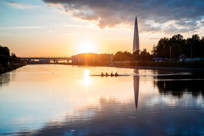 Boat regatta rowing team silhouette on the tranquil lake at sunset, cityscape on background.