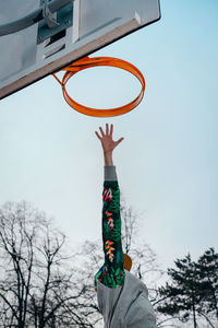 Low angle view of person with arm raised by basketball hoop against sky