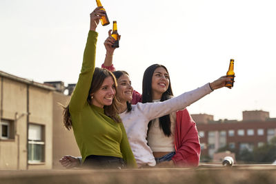 Happy female friends raising beer bottles enjoying on rooftop while looking away