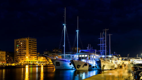 Sailboats moored on river by illuminated buildings against sky at night