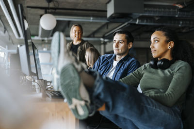 Female coder sitting with feet up sharing computer screen with male colleague while sitting in office