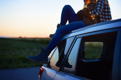 Man sitting in car against sky during sunset