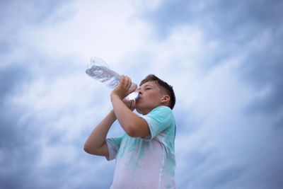 A boy against the sky drinks water from a plastic bottle. thirst. heat. cloudy sky.