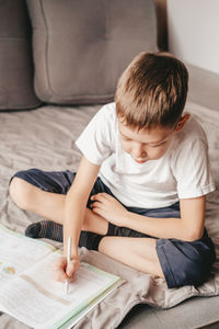 Boy does homework while sitting on the gray sofa. a caucasian teenager writes in a workbook