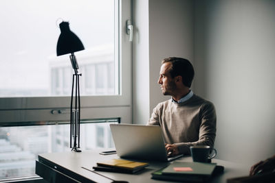 Thoughtful businessman looking through window while sitting with laptop at creative office