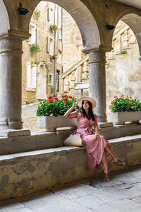 Young woman in pink dress sitting on stone wall of old building, town, picturesque.