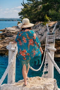 Rear view of young woman standing on old rope bridge over sea cove