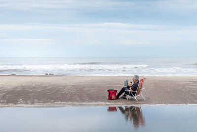 Mature man with gray hair and glasses sitting on a beach chair drinking mate and behind the waves