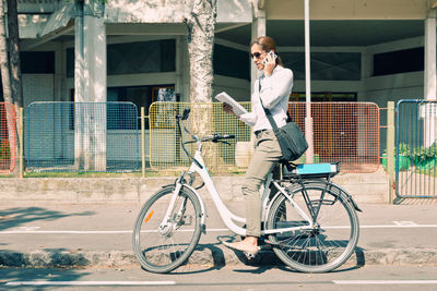 Side view of woman talking on smart phone while standing with bicycle on road