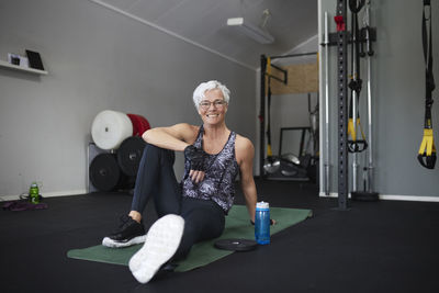 Full length portrait of smiling senior woman sitting on exercise mat at health club