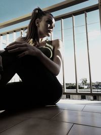 Young woman looking away while sitting on footpath by railing