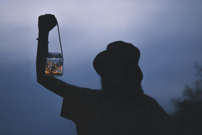 Silhouette of woman holding illuminated string lights in jar against blue sky