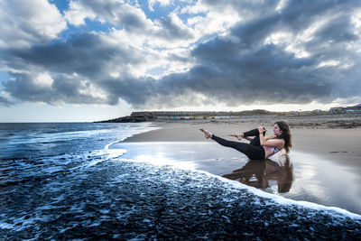 Young woman exercising while lying at beach against cloudy sky during sunset