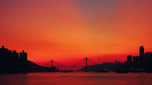 Silhouette bridge over river in city against orange sky