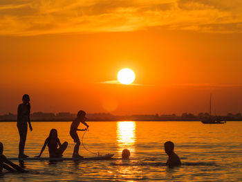 Silhouette people paddleboarding on sea against orange sky during sunset