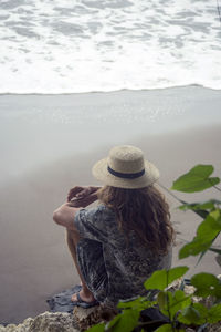 Indonesia, java, woman wearing straw hat sitting on rock on the beach looking to the sea